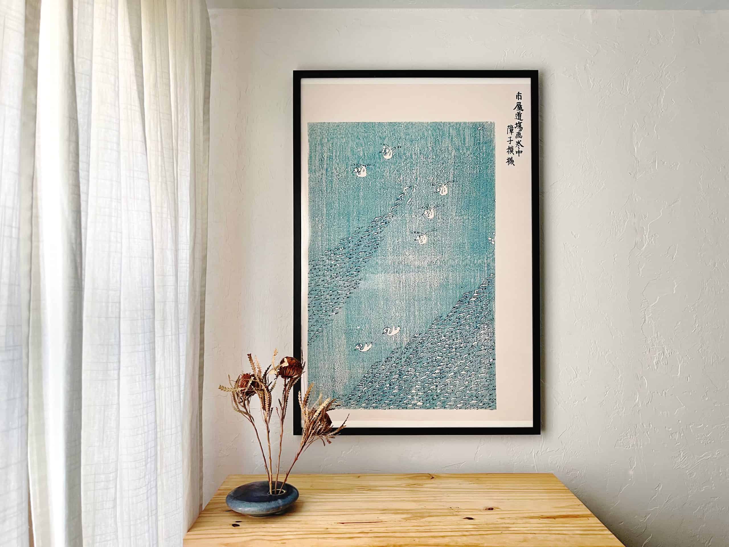 Frame poster of Blue lake by Taguchi Tomoki in black simple black frame hung above dresser with Japanese ikebana vase on one side, soft light entering through curtain for article from office posters to furniture: Unleash creativity with Society6